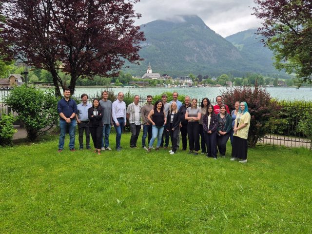 19 persons from the EXDIGIT team are standing on a meadow in front of a lake and mountains.