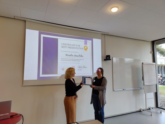 Nicola Leschke receives a certificate for the best presentation by chair member Laura Drechsler. In the background, the certificate is shown on a big screen.