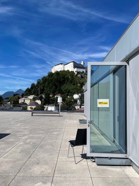 : On the right side of the picture, there is wall with a glass door, hold open through a chair, with a sign “PET-con”. The main part of the picture is a grey terrace, leading to the background picturing the fortress mountain of Salzburg, with a few houses along the hill and the fortress on top. The skye is blue.