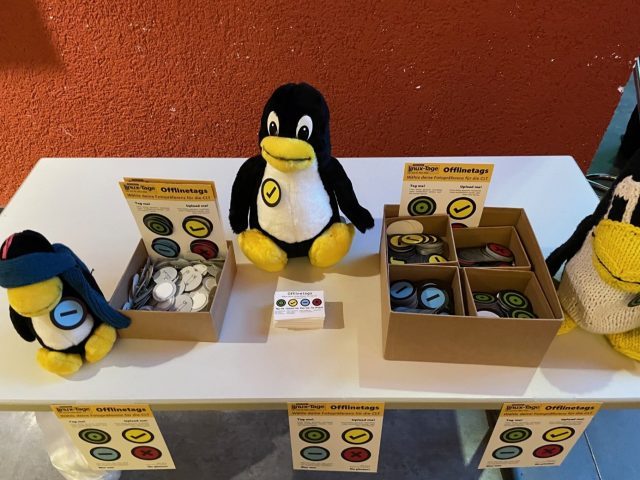 Ein Stand mit vielen Buttons mit den vier Offlinetagssymbolen, Informationsflyern und Visitenkarten. Dazwischen stehen drei Tuxe, jeweils mit einem Offlinetag.